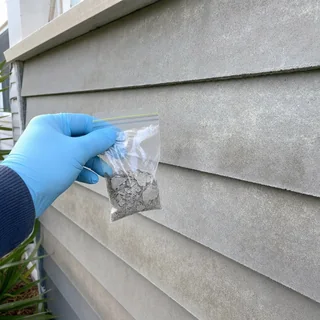 Gloved hand taking a small sample beside an old fibre cement sheet wall